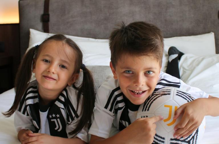Due bambini sorridenti con maglie della Juventus e un pallone su un letto.